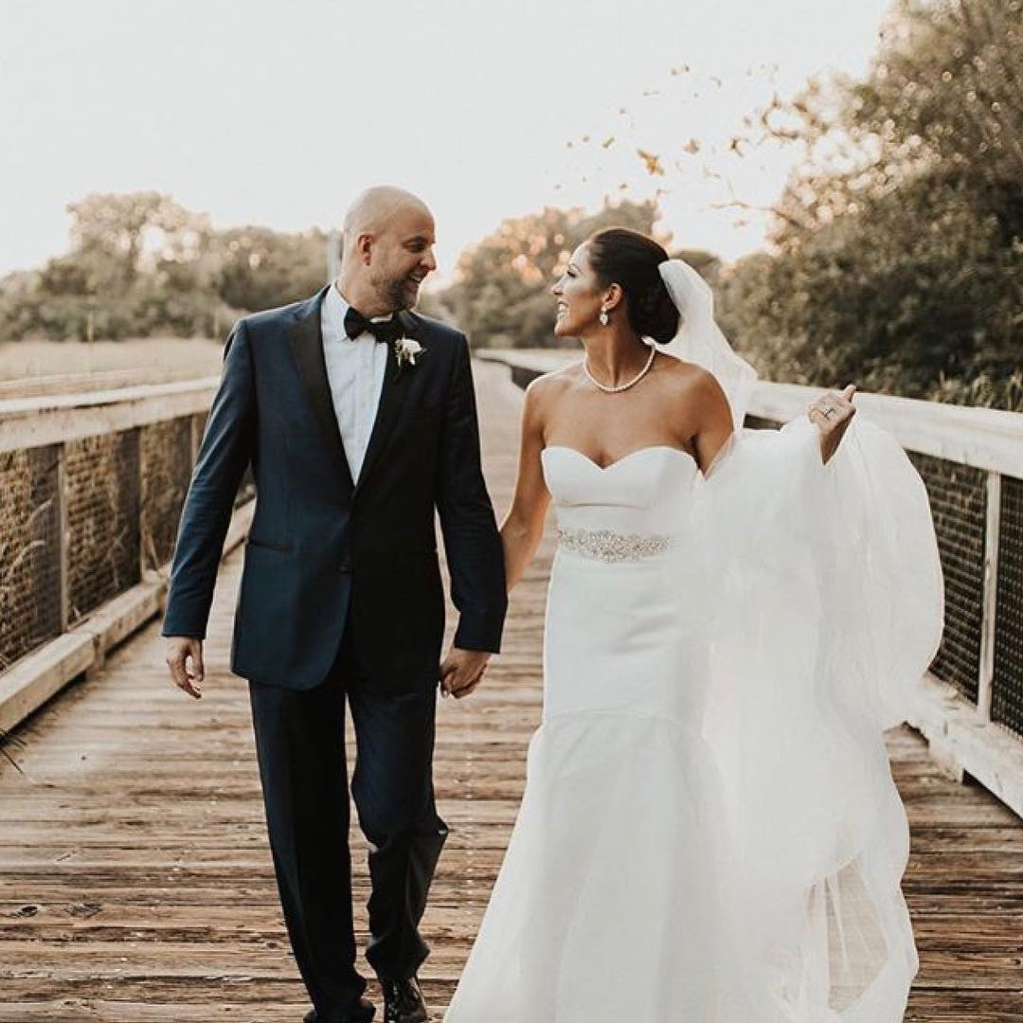 Bride and groom walking hand-in-hand, smiling, in custom wedding attire on a wooden bridge at sunset.