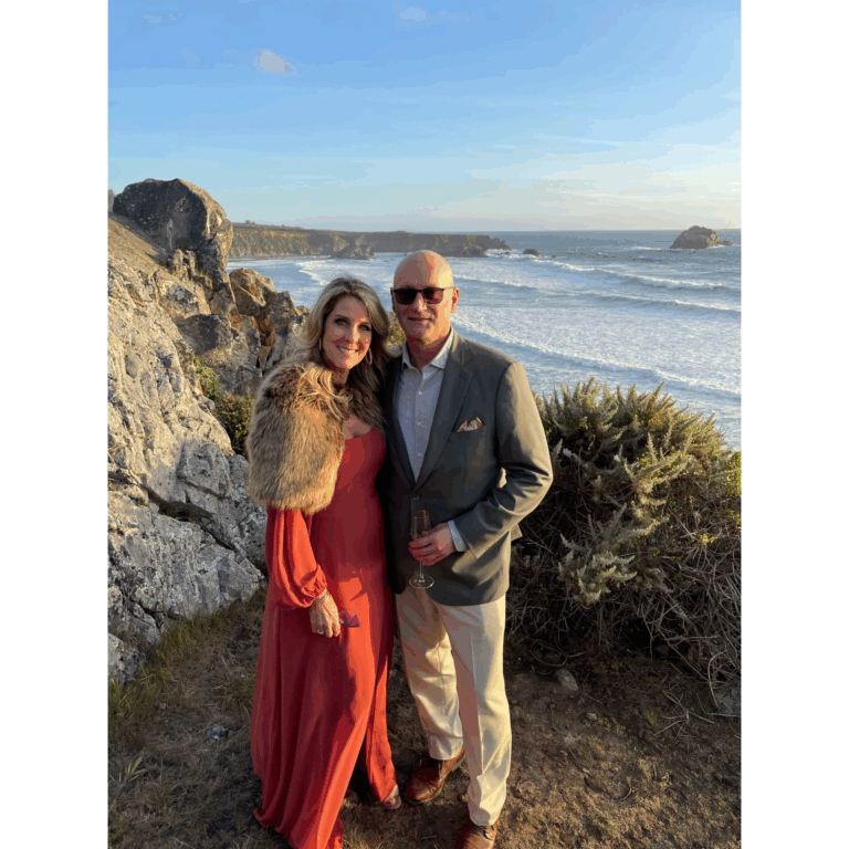 Couple dressed in elegant evening wear, standing by the ocean at sunset—man in a custom sport coat and woman in a rust maxi dress with fur shawl.