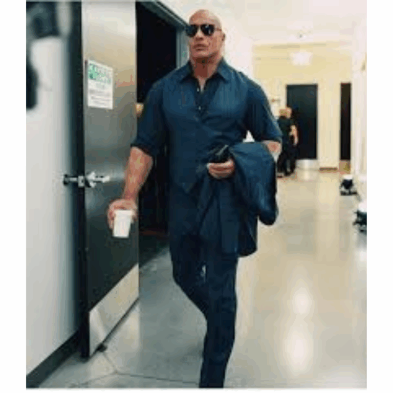 Dwayne “The Rock” Johnson walking confidently through a backstage hallway wearing a custom navy J.Hilburn shirt and pants, holding a coffee.