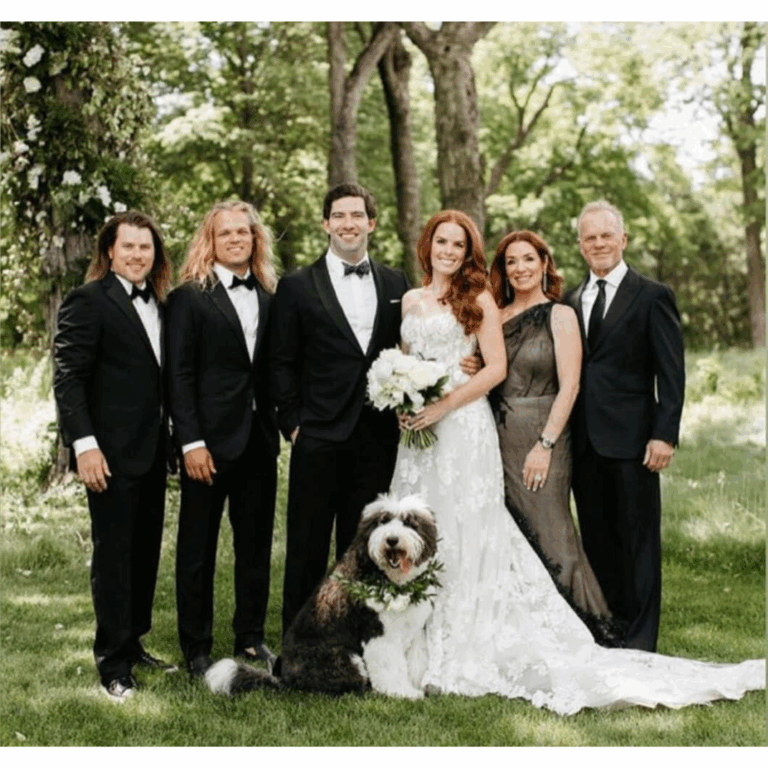 Outdoor wedding family portrait featuring bride and groom, three men in custom tuxedos, two women in formal gowns, and a floral-collared dog.