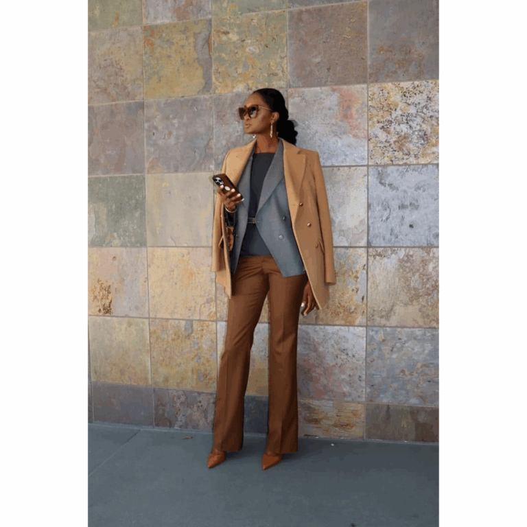 Stylish woman in a camel coat draped over a gray blazer, brown trousers, and pointed heels, standing confidently against a stone wall.