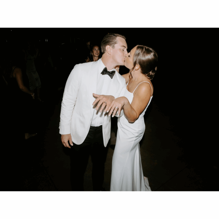 Bride and groom sharing a kiss at their wedding reception; bride showing her ring, groom in a white tuxedo jacket and black bow tie.