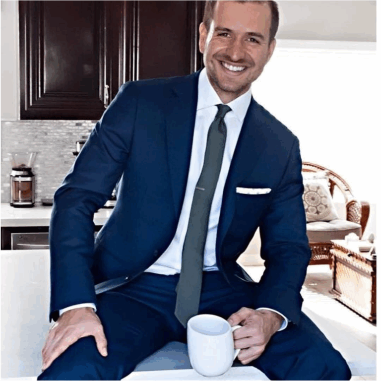 Smiling man in a navy blue custom suit and tie, sitting casually on a kitchen counter holding a white coffee mug.