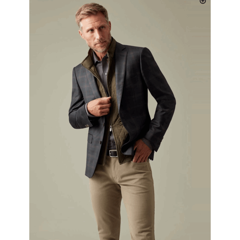 J.Hilburn male model in layered fall outfit—olive quilted vest under a dark plaid sport coat, with tan trousers and dress shirt.