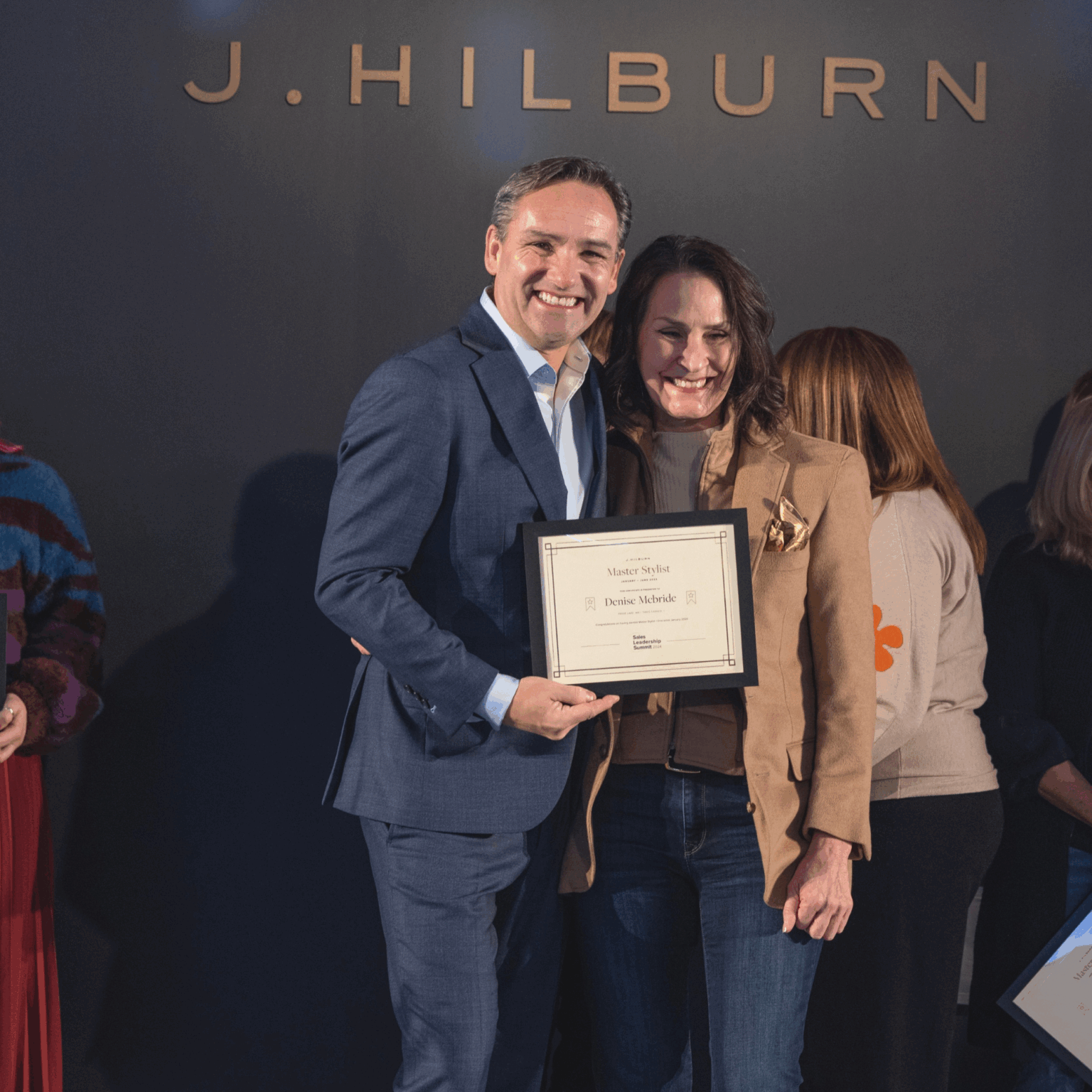Denise McBride receiving the J.Hilburn Master Stylist award on stage, smiling with executive presenter.